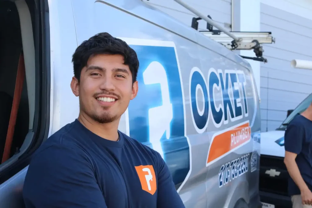 A smiling young man with dark hair and a small beard, wearing a navy blue "Pocket Plumber" t-shirt, stands in front of a service van